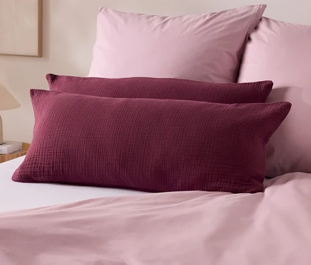Deux coussins bordeaux sont posés sur un lit avec des draps roses. À l'arrière-plan, deux coussins roses et une lampe.