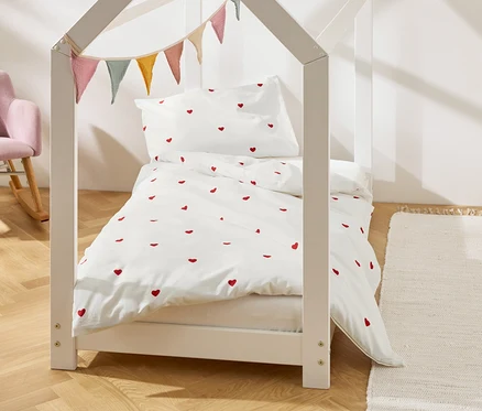Un lit d'enfant en forme de maison avec des draps blancs à motifs de cœurs rouges et des fanions. Un fauteuil rose est visible en arrière-plan.