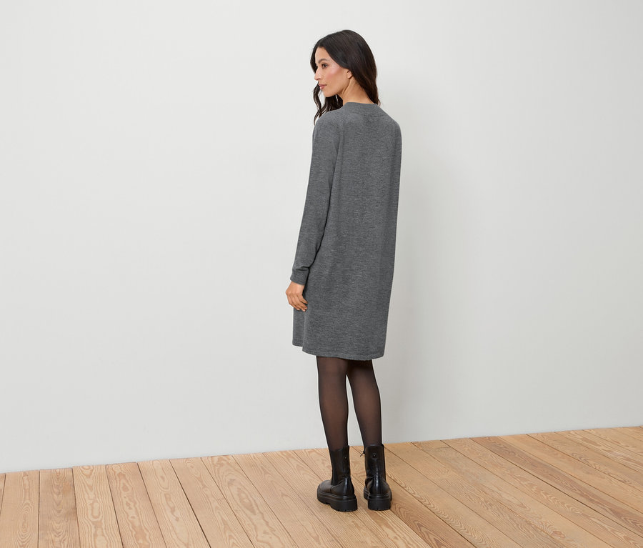 Une femme se tient dos à la caméra, portant une robe grise, des collants noirs et des bottes noires.