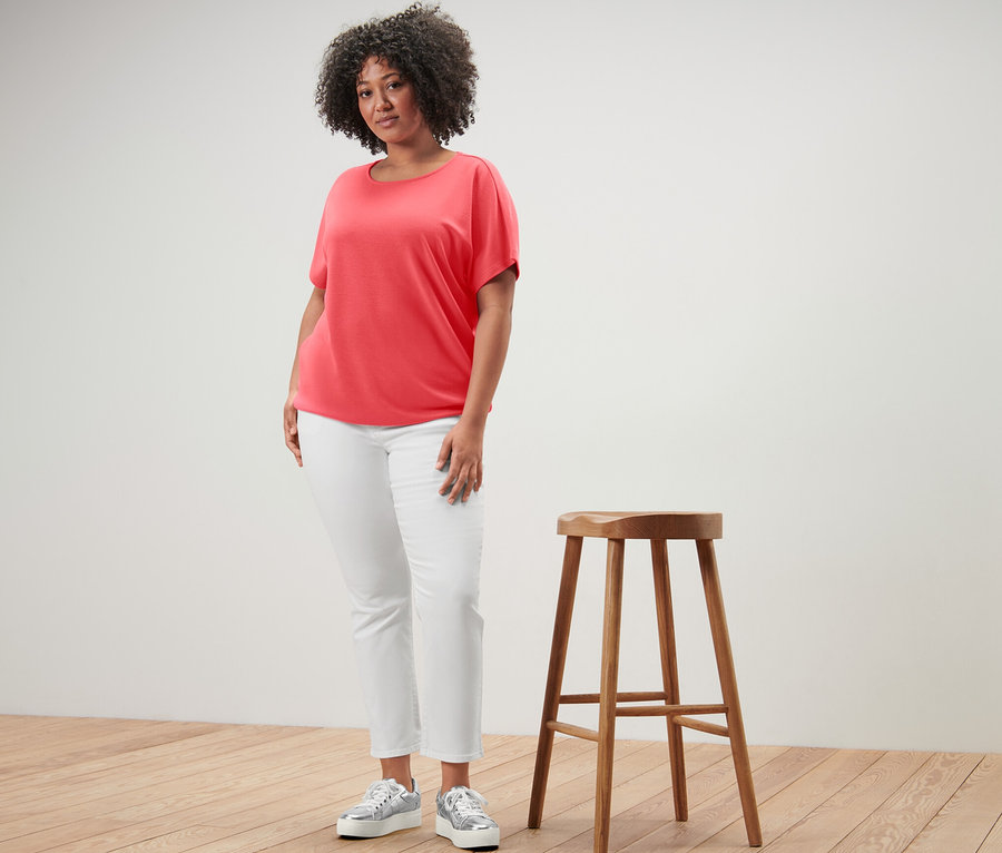 Une femme debout à côté d'un tabouret en bois porte une blouse corail.