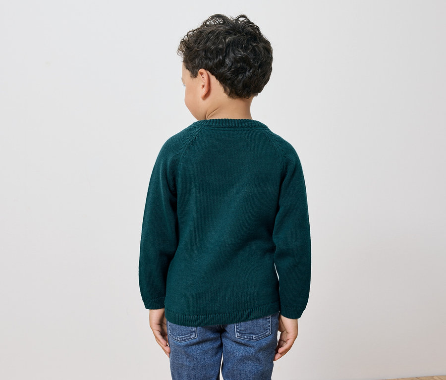 Un garçon aux cheveux bouclés et foncés se tient dos à la caméra, portant un pull vert et un jean.