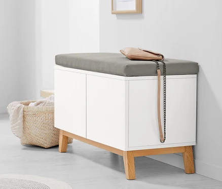 Un banc blanc avec des étagères de rangement et un coussin gris. Un sac à main beige est posé sur le banc.
