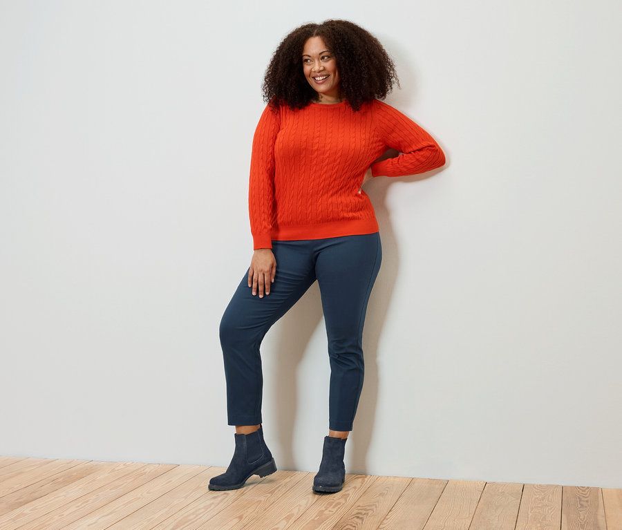 Femme souriante aux cheveux bouclés posant en pull orange, pantalon bleu et bottes.