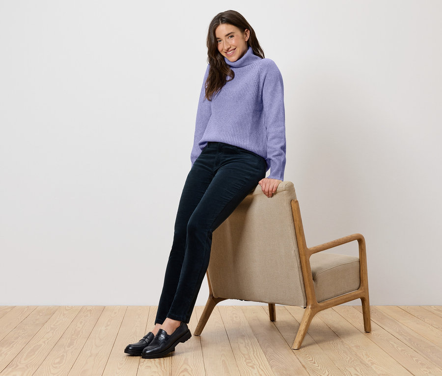 Une femme est assise sur une chaise beige, portant un pull à col roulé violet, un pantalon bleu marine et des chaussures noires.