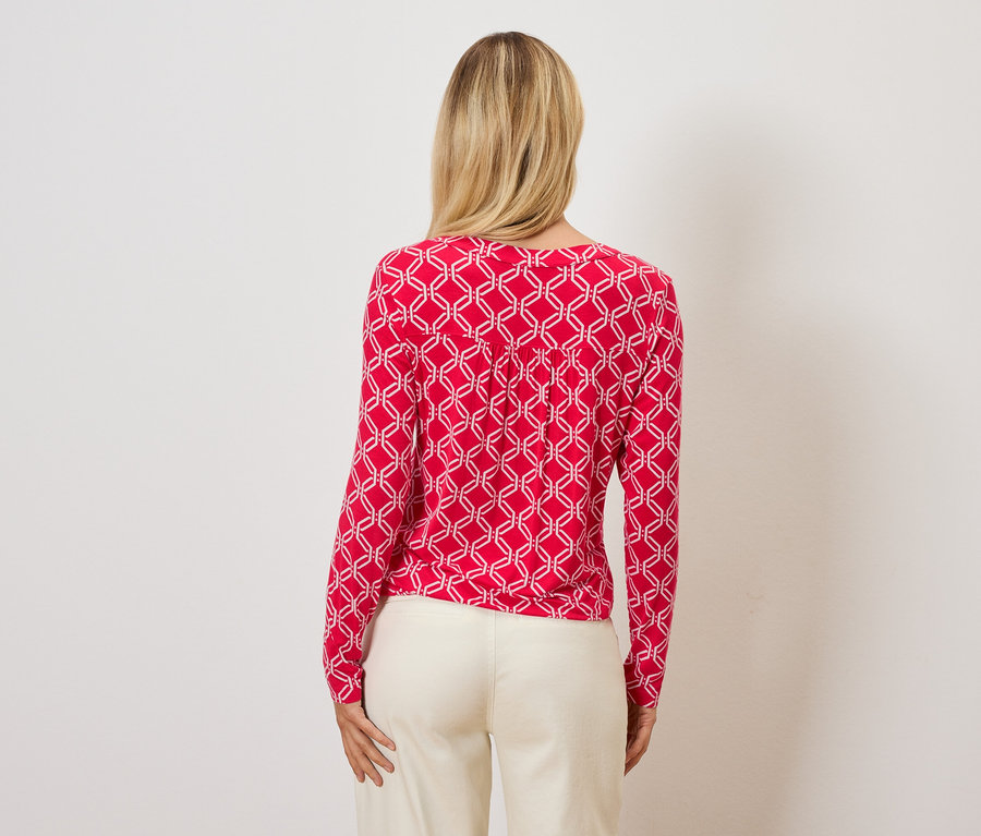 Vue de dos, une femme aux cheveux blonds porte un haut rouge avec un motif géométrique blanc et un pantalon crème.