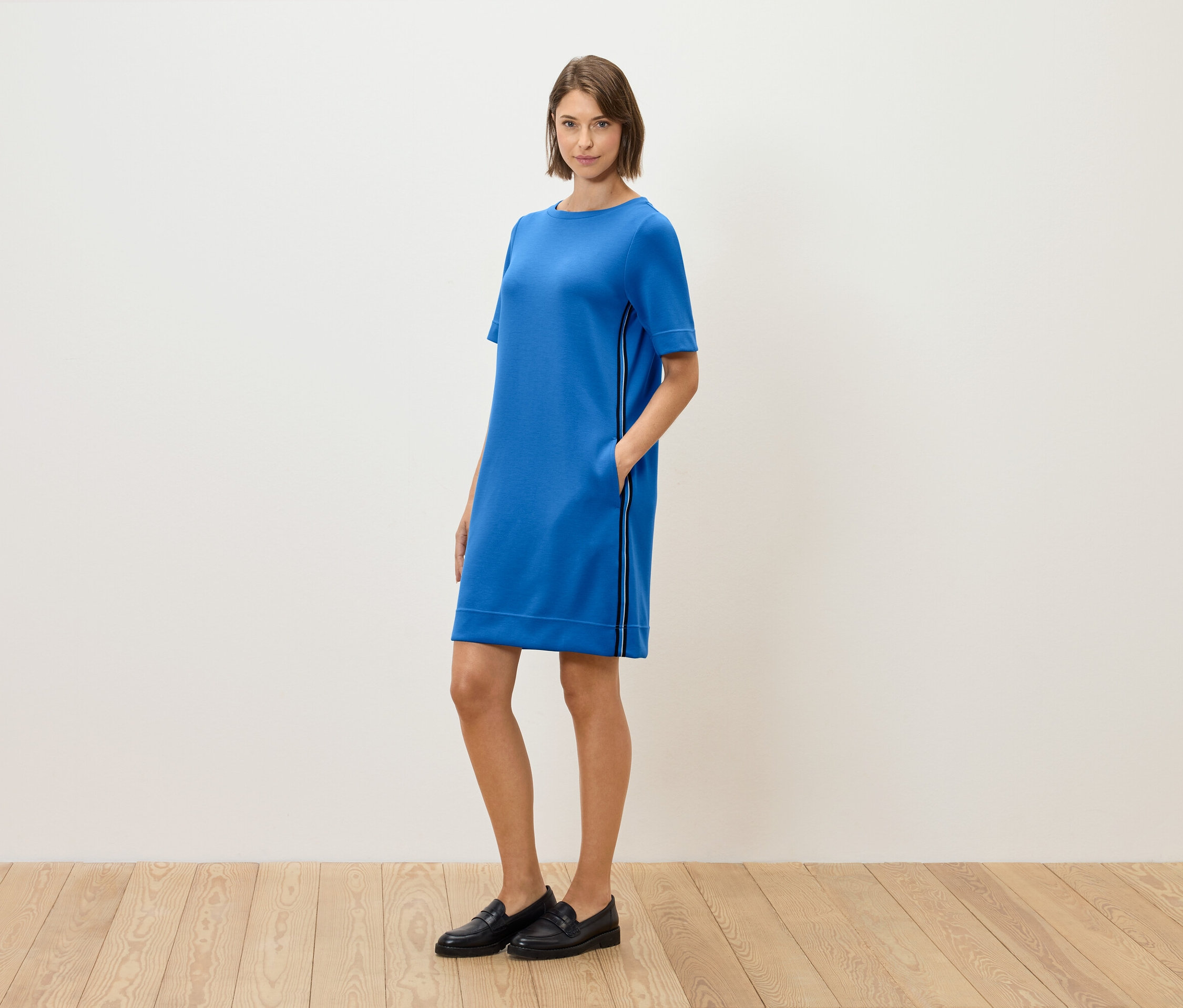 Une femme debout, pose dans une robe bleue à manches courtes et des mocassins noirs.