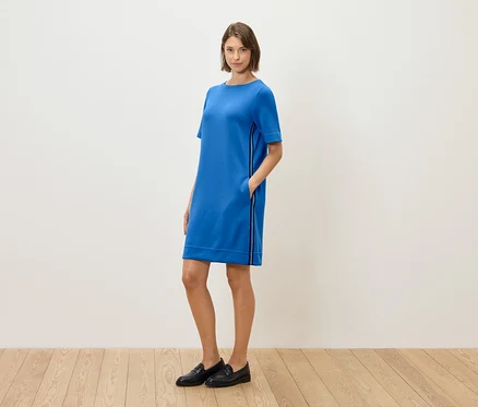 Une femme debout, pose dans une robe bleue à manches courtes et des mocassins noirs.