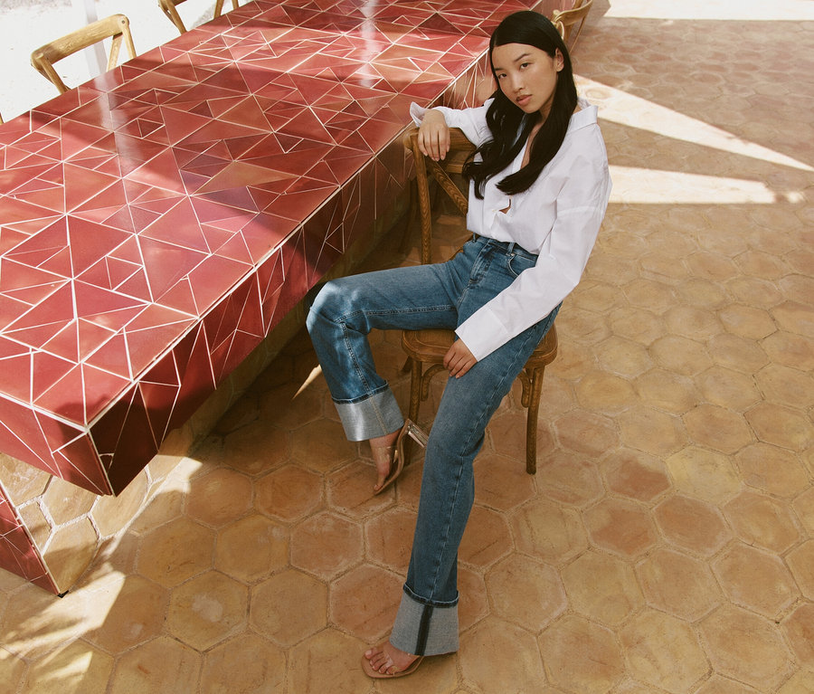 Une femme assise sur une chaise à une table porte un jean Angels « Lara » coupe droite avec revers, bleu clair.