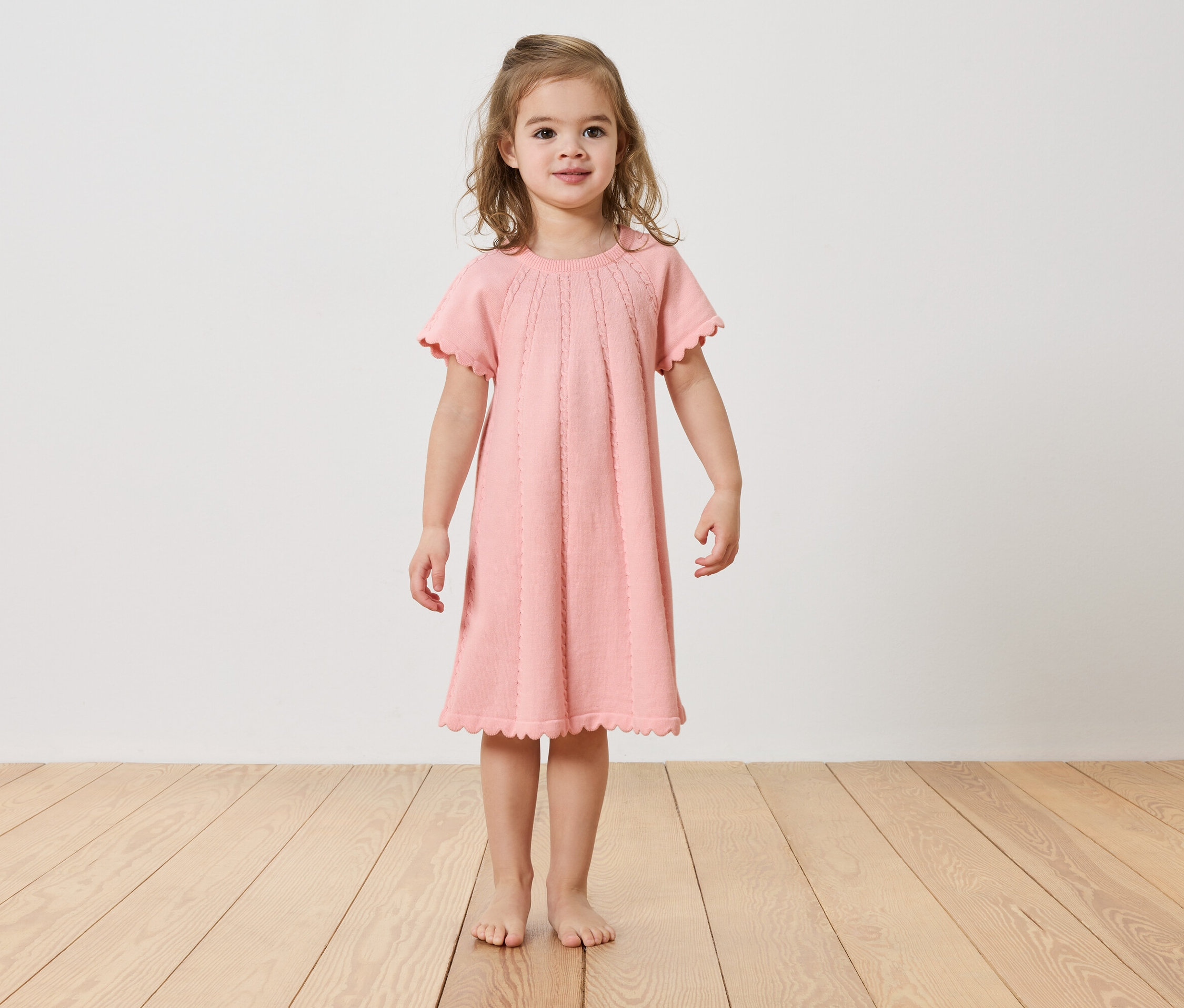 Fillette debout pieds nus sur un plancher en bois, portant une robe rose.