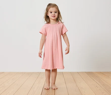 Fillette debout pieds nus sur un plancher en bois, portant une robe rose.