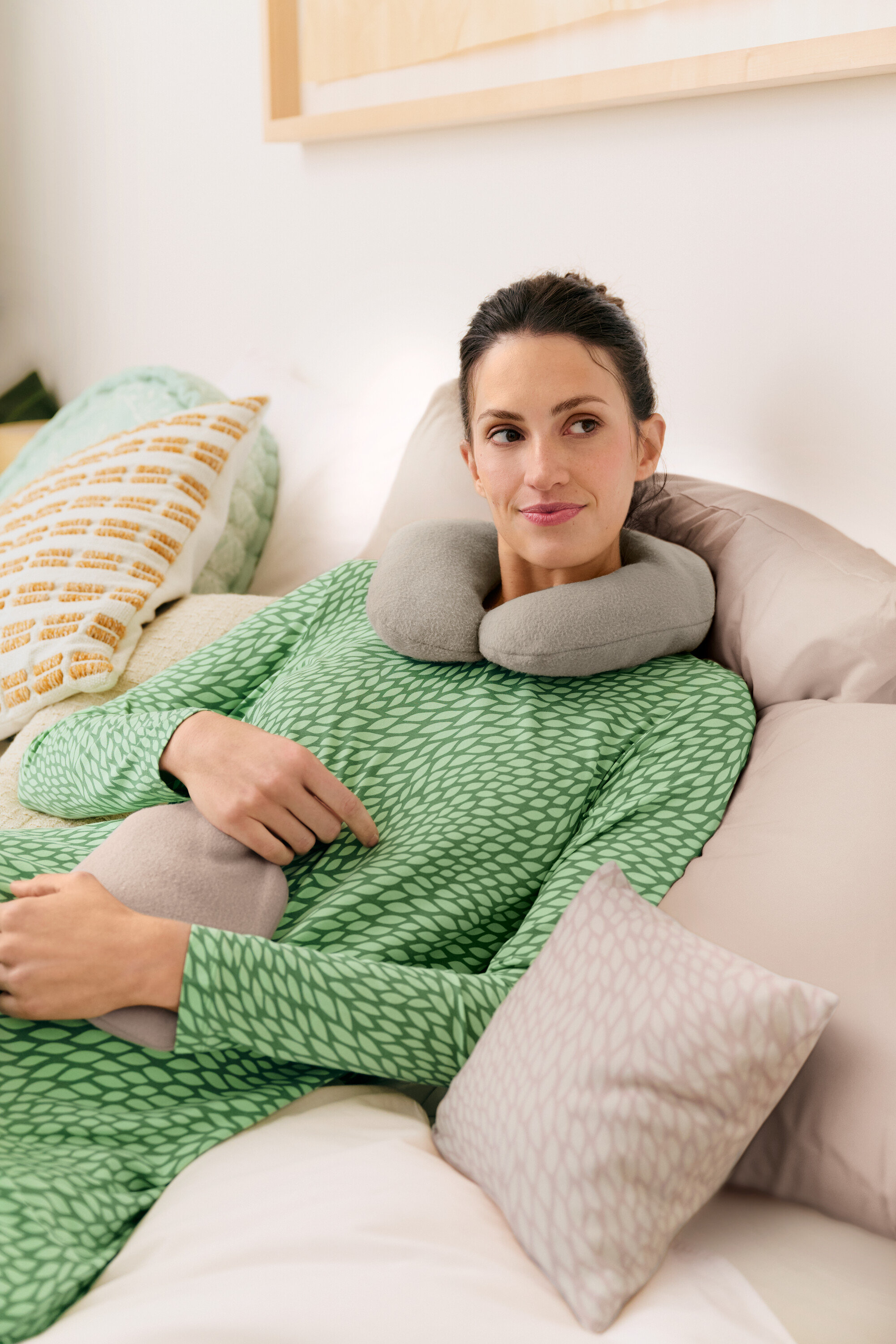 Femme allongée dans un lit avec un coussin de voyage gris et une bouillotte, portant une chemise de nuit verte.