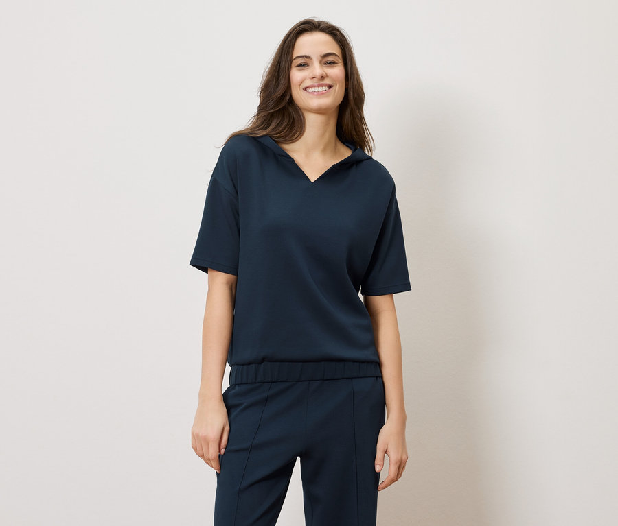 Une femme brune souriante porte un ensemble bleu marine : un haut à manches courtes et un pantalon.