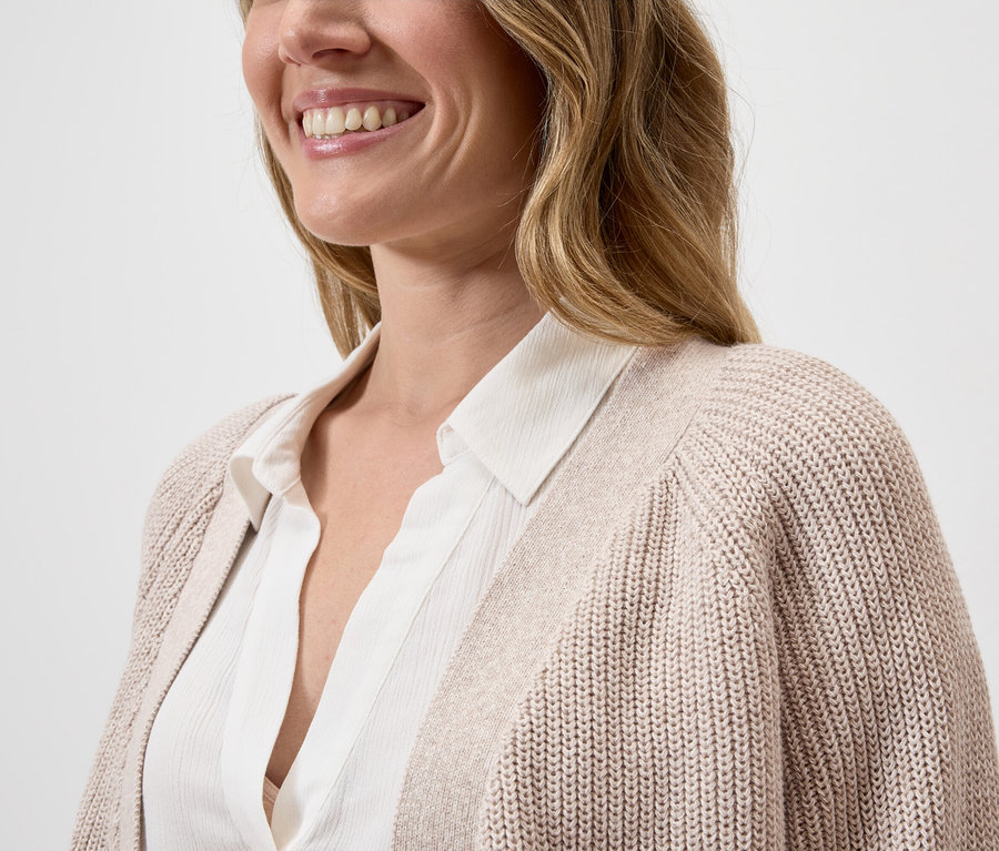 Une femme souriante porte un chemisier blanc sous un cardigan beige.