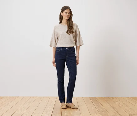 Une femme portant un pull beige et un jean foncé se tient sur un plancher en bois devant un mur blanc.
