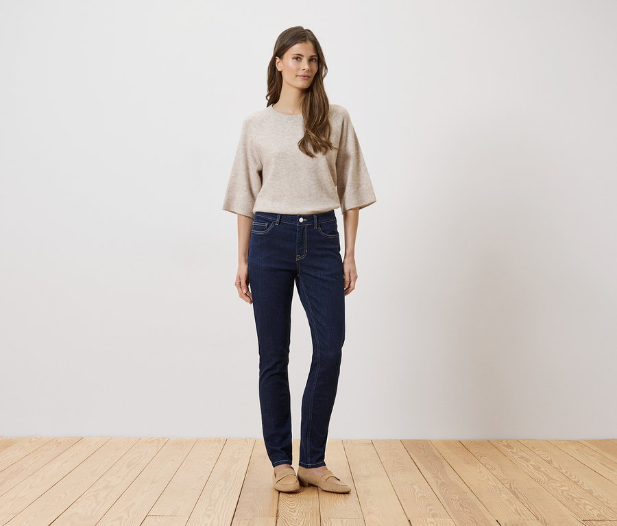 Une femme portant un pull beige et un jean foncé se tient sur un plancher en bois devant un mur blanc.
