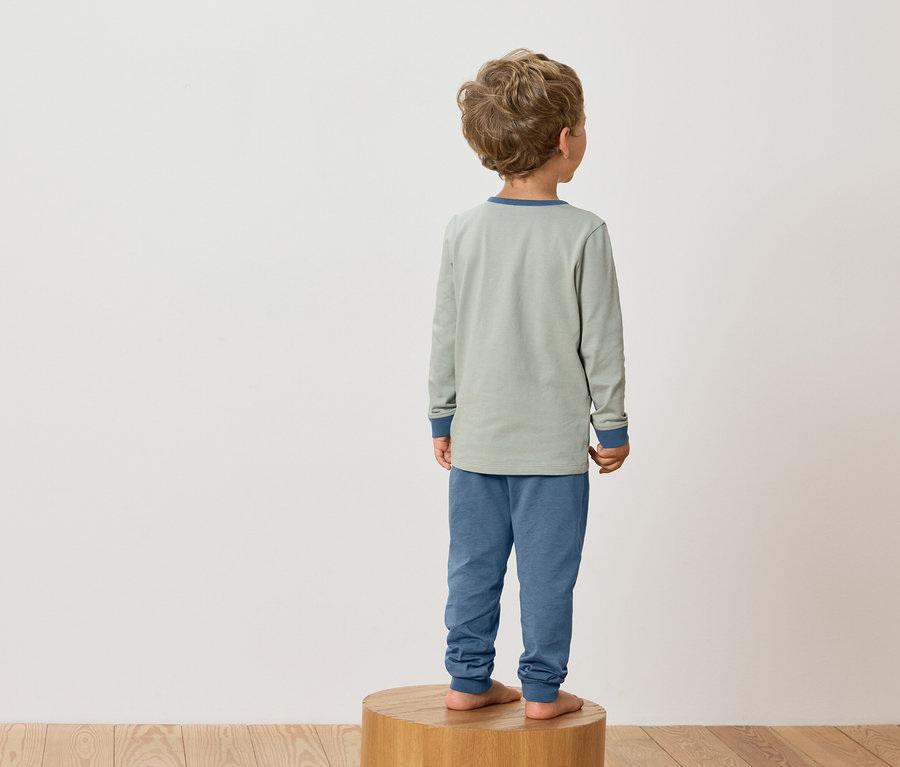 Un enfant aux boucles blondes se tient pieds nus sur un tabouret en bois et porte un pyjama vert avec des poignets bleus.
