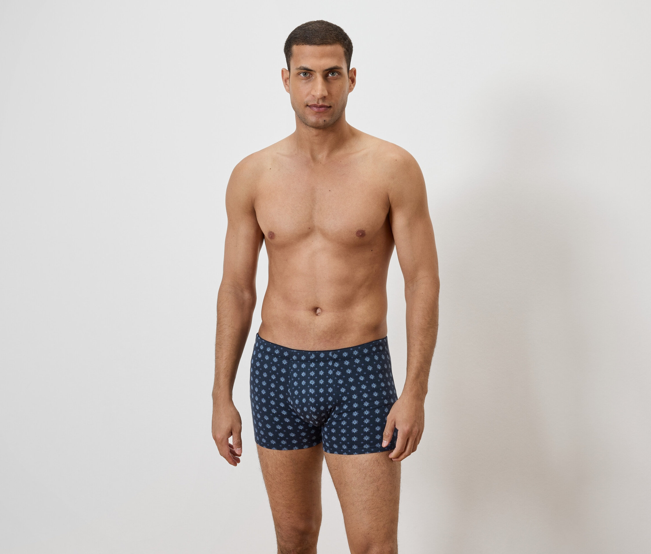 Un homme torse nu se tient devant un mur blanc et porte un caleçon boxeur bleu avec un motif floral répété.