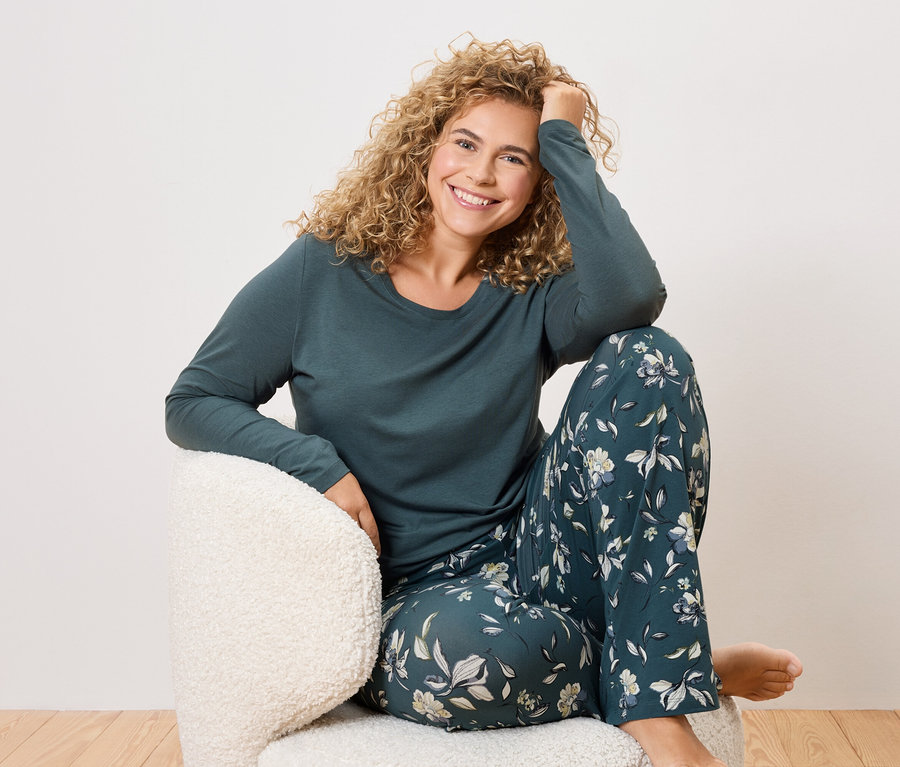 Une femme aux cheveux bouclés est assise dans un fauteuil et porte un pyjama vert foncé à motifs floraux.