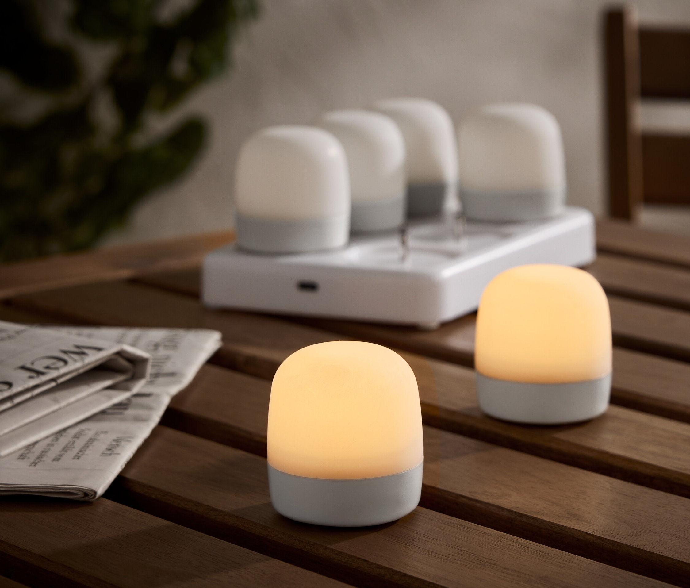 Deux lampes de table sans fil illuminées sur une table en bois, avec une station de charge et quatre lampes supplémentaires en arrière-plan. Un journal est posé à côté.