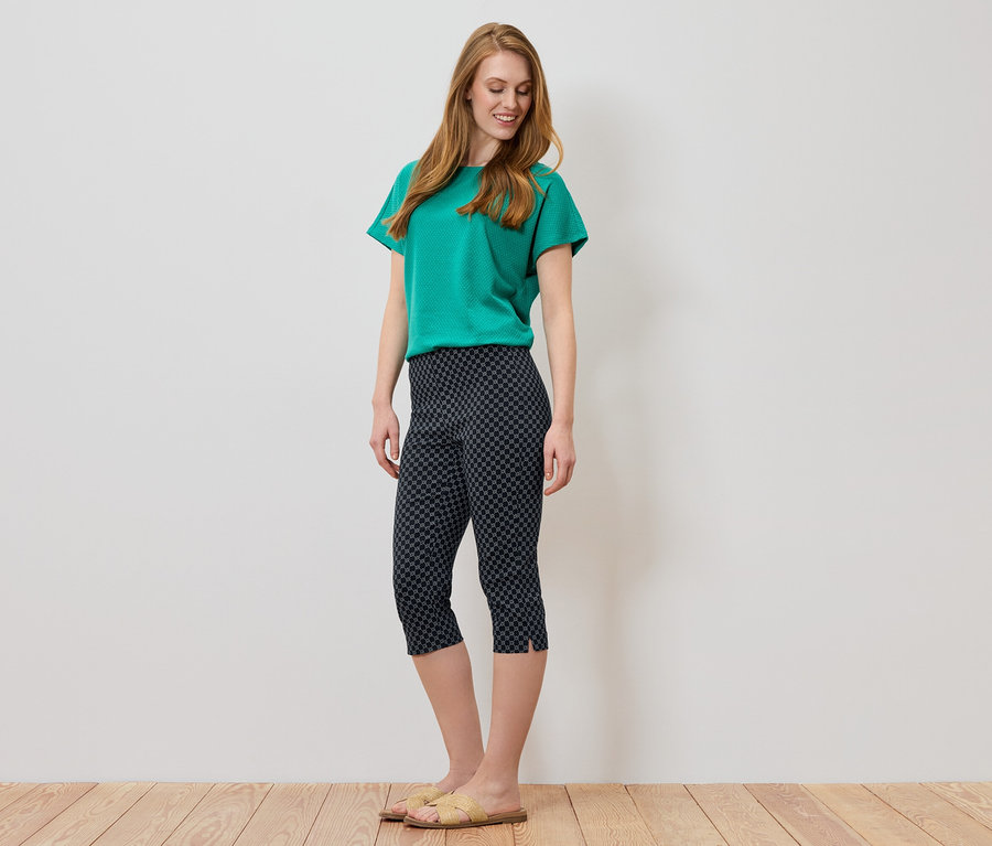 Femme aux longs cheveux roux portant un t-shirt vert et un pantacourt foncé à motifs géométriques.