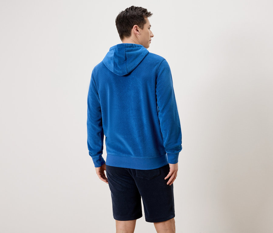 Homme vu de dos portant un sweat à capuche bleu en tissu éponge et un short devant un fond neutre.