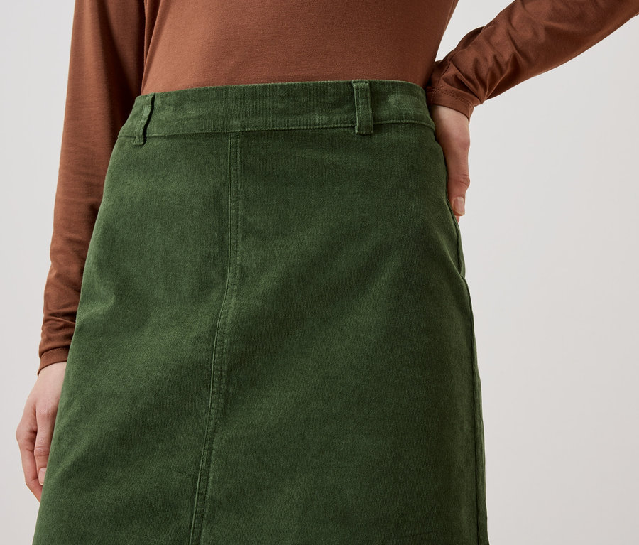 Gros plan d'une femme portant un haut marron et une jupe en velours côtelé vert olive, la main sur la hanche.