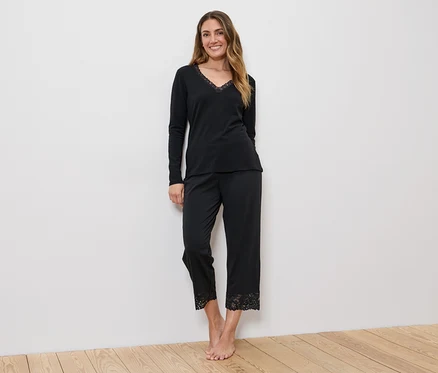 Femme portant un haut noir à manches longues et col en V et un pantalon noir avec de la dentelle en bas, devant un mur blanc.