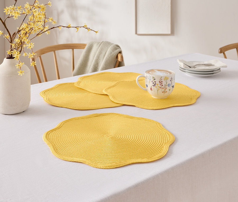 Sur une table avec une nappe blanche se trouvent un vase avec des fleurs jaunes, trois sets de table jaunes et une tasse à motif floral.