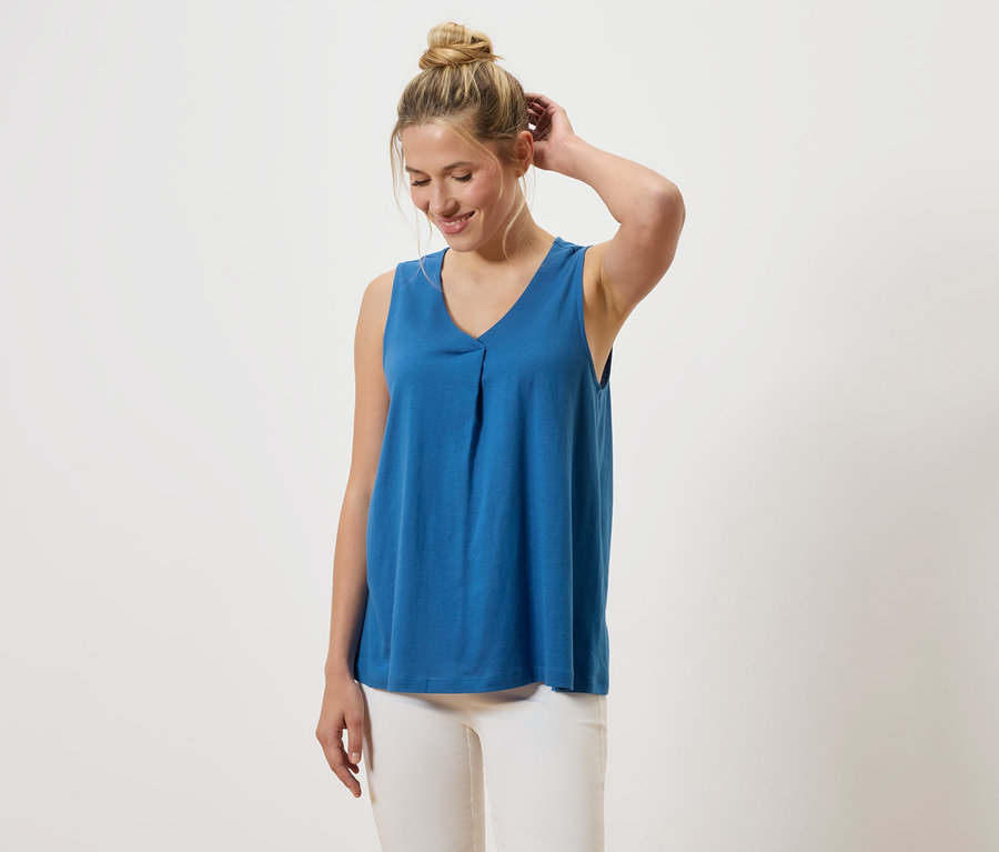 Une femme aux cheveux blonds porte un haut bleu à col en V et un pantalon blanc.