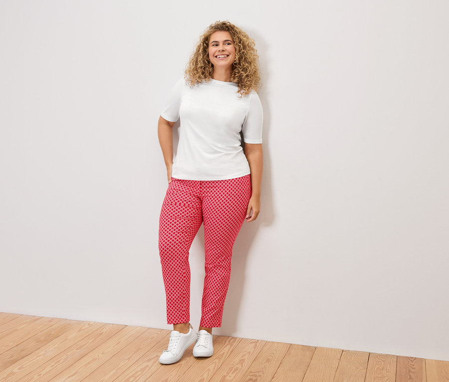 Une femme aux cheveux bouclés, portant un t-shirt blanc, un pantalon à motifs rouges et des baskets blanches, se tient devant un mur blanc.