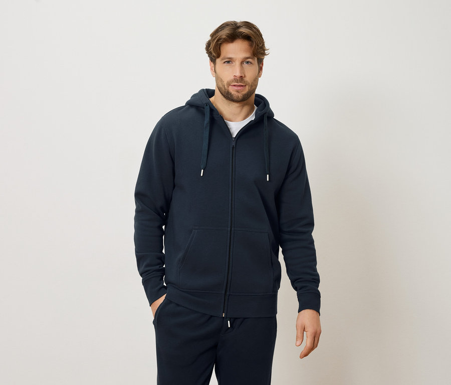 Homme portant un sweat à capuche zippé bleu marine et un pantalon de survêtement bleu marine, debout devant un fond crème.