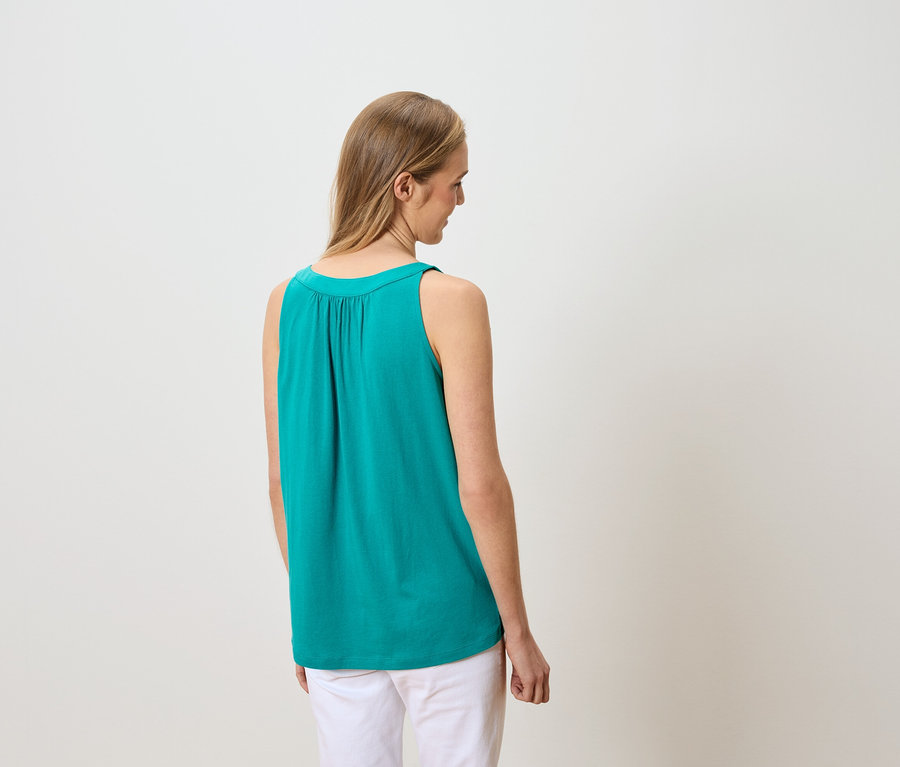 Vue arrière d'une femme blonde portant un débardeur turquoise et un pantalon blanc.