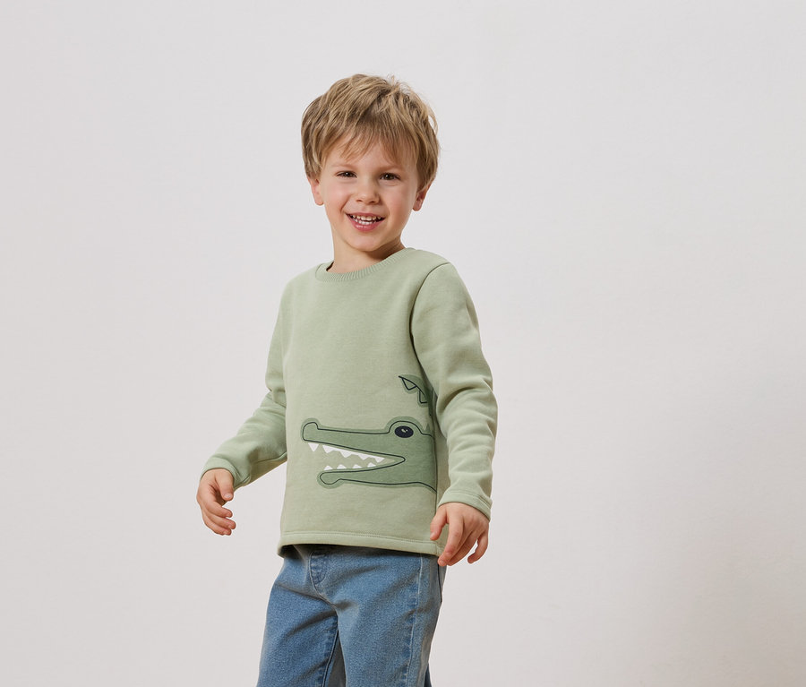 Un garçon souriant porte un sweat-shirt vert clair avec un crocodile dessus et un jean.