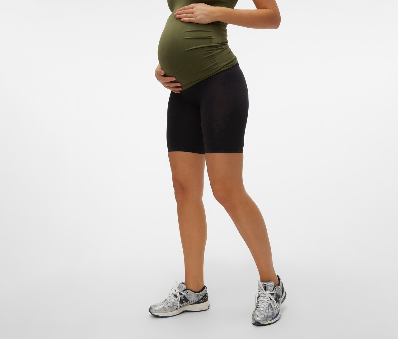 Femme enceinte portant un haut olive et un Skinny Shorts MAMALICIOUS «
MLTIA JEANNE
», mellow buff noir, les mains sur le ventre, avec des baskets.