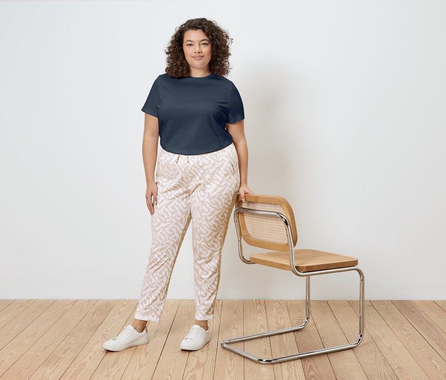 Une femme aux cheveux bouclés se tient à côté d'une chaise avec un cadre en bois et une assise en rotin. Elle porte un t-shirt bleu marine, un pantalon à motifs et des baskets blanches.