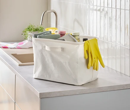 Sur un comptoir de cuisine, une boîte en tissu blanc contient divers articles de nettoyage et des gants en caoutchouc jaunes.