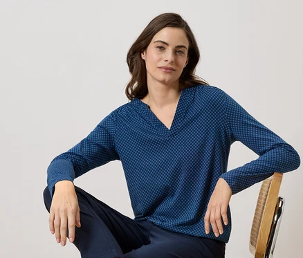 Femme assise sur une chaise en bois portant un chemisier bleu marine à manches longues avec un motif à pois et un pantalon bleu marine.