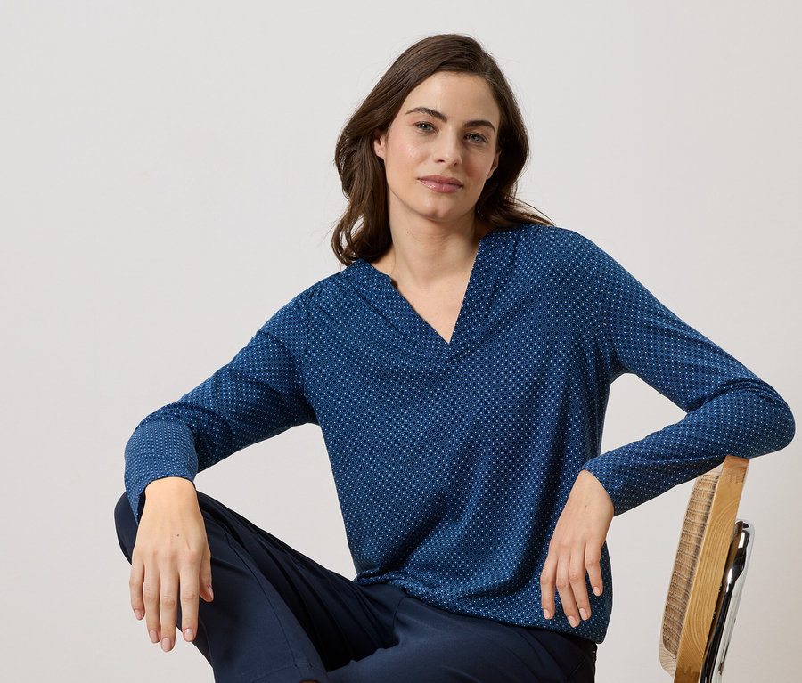 Femme assise sur une chaise en bois portant un chemisier bleu marine à manches longues avec un motif à pois et un pantalon bleu marine.
