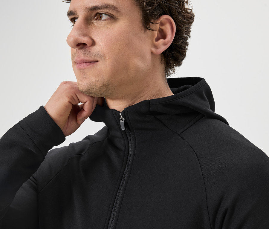 Un homme aux cheveux bouclés porte un sweat à capuche noir zippé et tient une main sur le col.