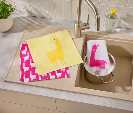Un torchon jaune et un torchon rose à motif de lama sont posés à côté d'un évier dans lequel se trouve une casserole avec un torchon à motif de lama.