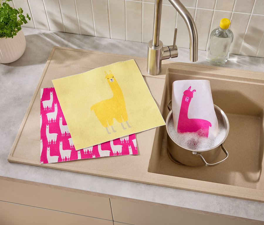 Un torchon jaune et un torchon rose à motif de lama sont posés à côté d'un évier dans lequel se trouve une casserole avec un torchon à motif de lama.