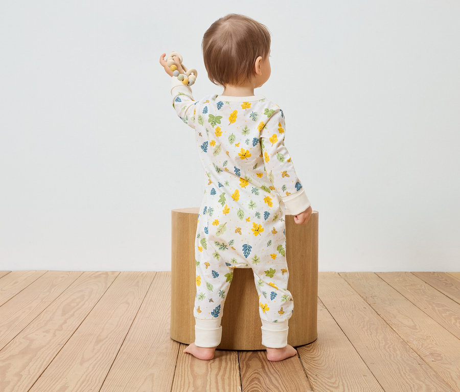 Un bébé en combinaison à motifs de feuilles se tient dos à un tabouret en bois et tient un jouet en bois.