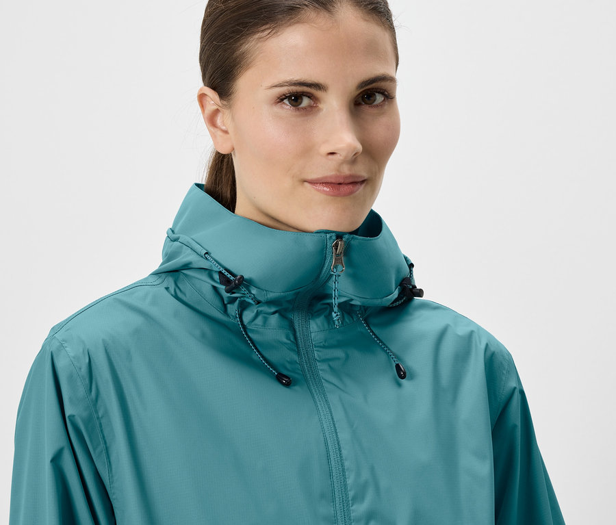 Une femme avec une queue de cheval porte un imperméable turquoise avec une capuche et une fermeture éclair sur fond blanc.