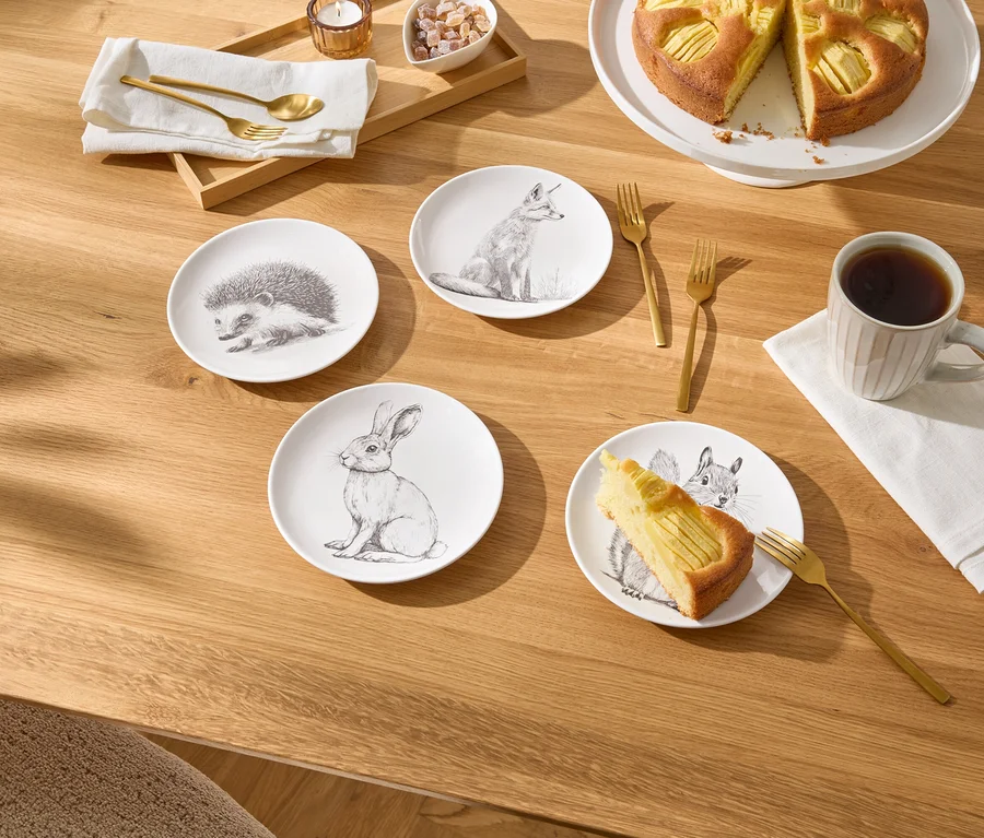 Sur une table en bois, il y a quatre petites assiettes avec des dessins d'animaux, une part de gâteau aux pommes, une tasse de café et des couverts dorés.