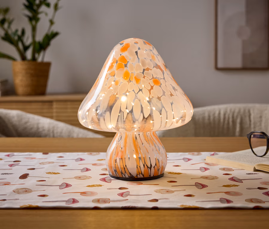 Une lampe en forme de champignon en verre moucheté orange et blanc avec des lumières internes est posée sur une table en bois. Un chemin de table blanc avec des motifs de champignons est placé en dessous. À droite, un livre ouvert avec des lunettes à monture foncée repose sur ses pages. Une plante en pot et un tableau encadré sont visibles en arrière-plan flou.