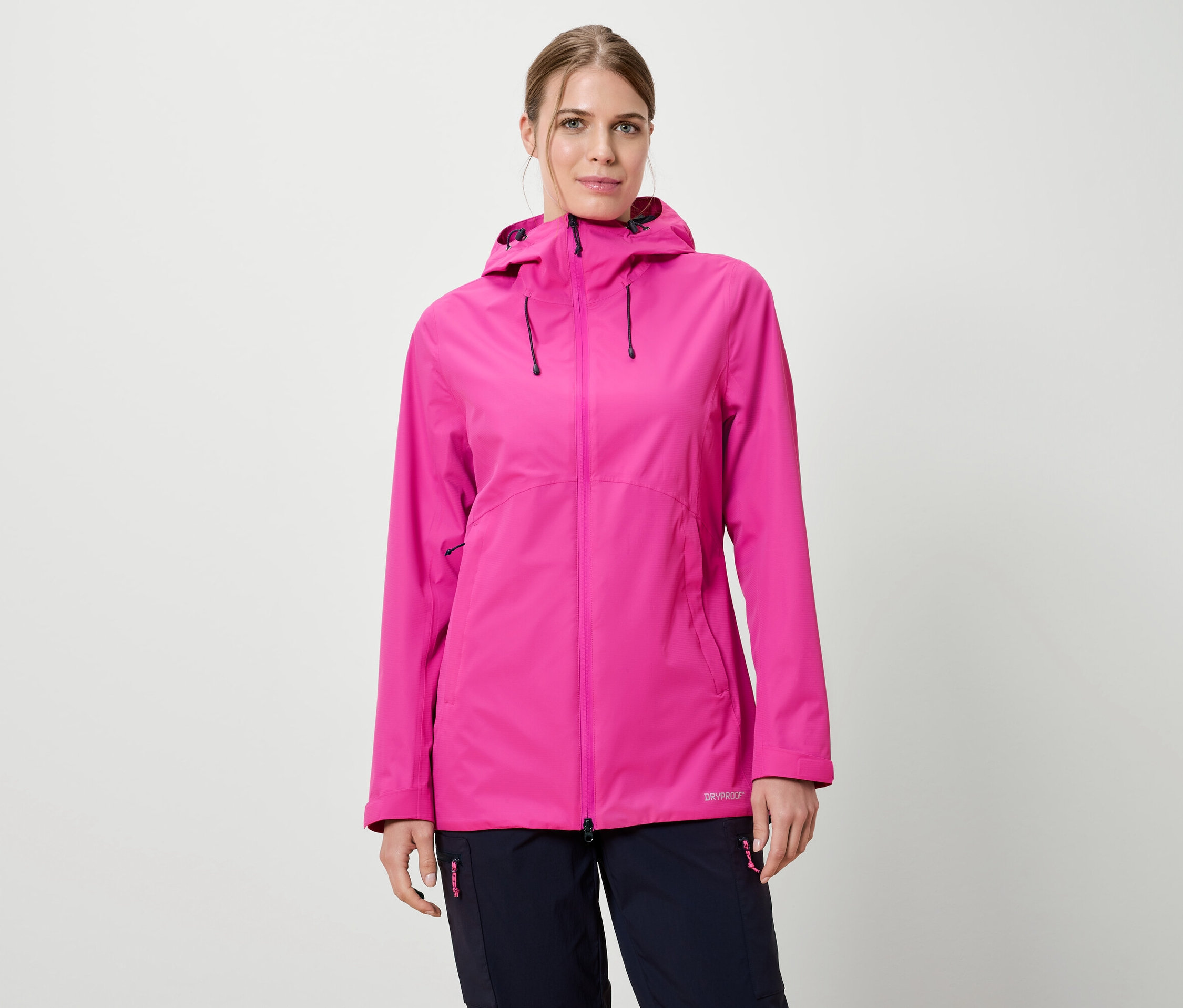 Une femme portant une veste de pluie rose à capuche et un pantalon bleu marine sur un fond blanc.