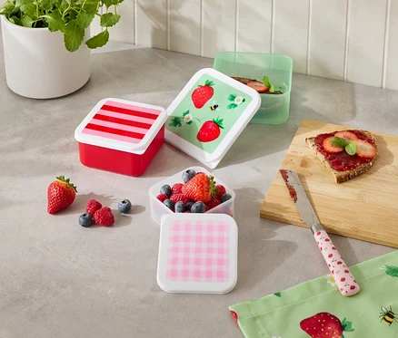Plusieurs boîtes de conservation colorées sont posées sur un comptoir de cuisine, certaines remplies de fruits, à côté d'une tranche de pain et d'un couteau.