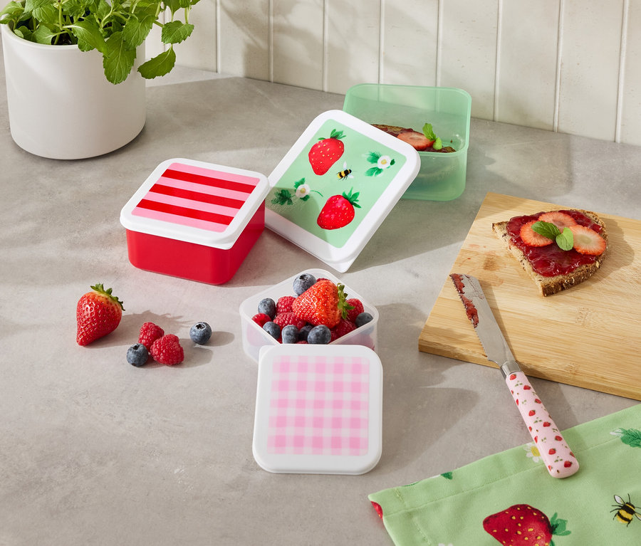 Plusieurs boîtes de conservation colorées sont posées sur un comptoir de cuisine, certaines remplies de fruits, à côté d'une tranche de pain et d'un couteau.