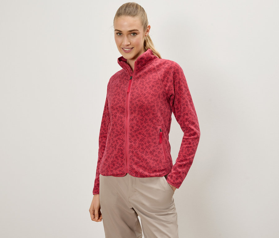 Femme posant en veste polaire rouge à fleurs et pantalon beige, une main dans la poche.