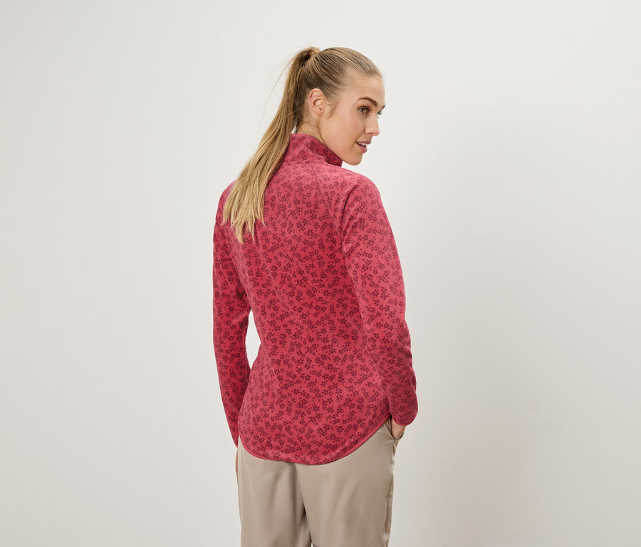 Femme blonde avec queue de cheval, vue de dos, portant un haut rouge à manches longues avec un motif floral et un pantalon beige.
